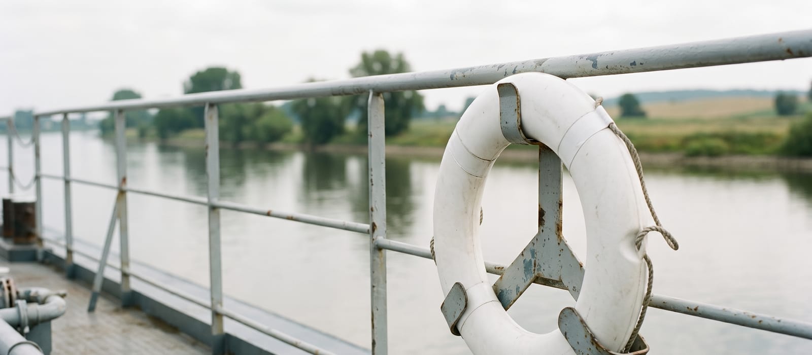 A white lifebuoy mounted on the railing of an inland barge, the river soft in the background.
