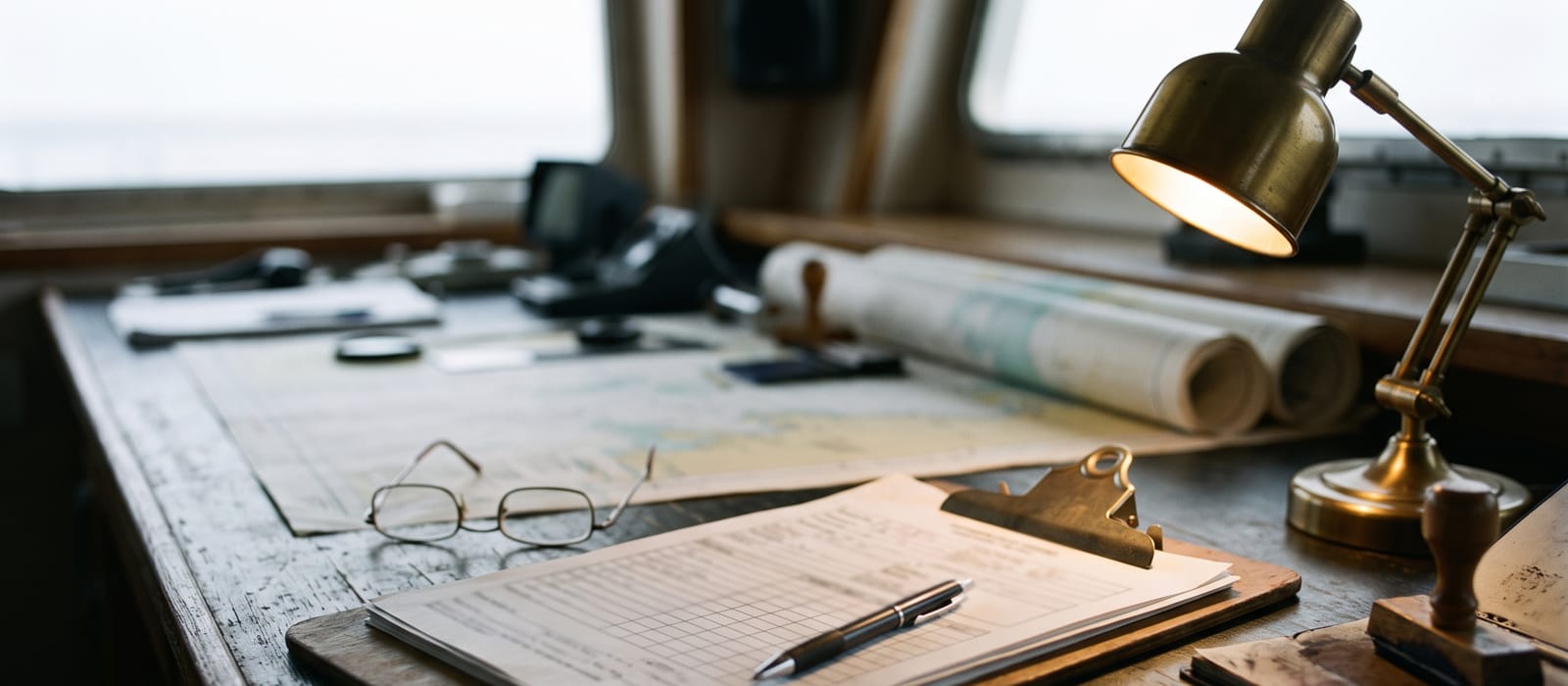 An inspection clipboard and pen on a chart table beside a stamp and reading glasses, paperwork out of focus.