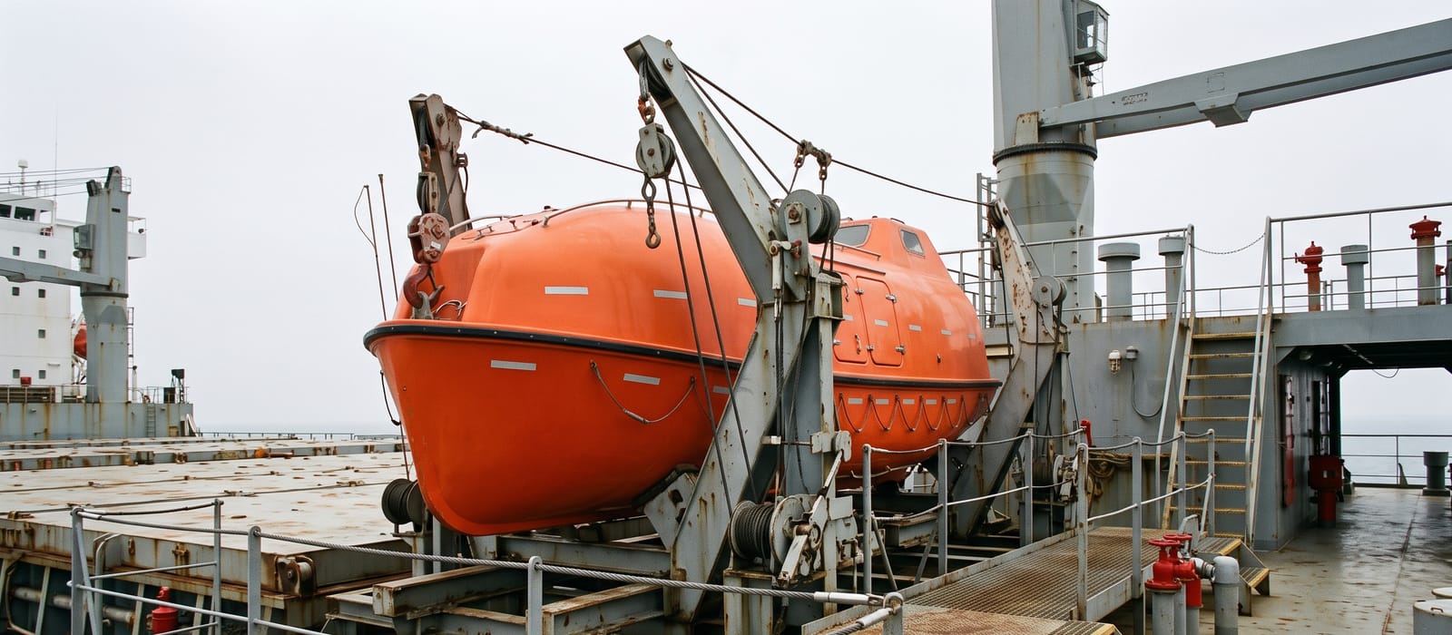 An orange enclosed lifeboat on its davits on a cargo ship's deck against a grey sky.