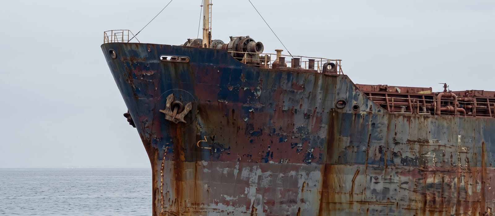 The weathered bow of an older bulk carrier at anchor, rust streaks running down the faded hull.